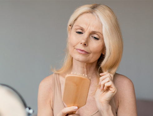 A imagem mostra um mulher loira de meia-idade olhando preocupada para uma escova de cabelo em suas mãos, refletindo os efeitos da menopausa na saúde dos cabelos.