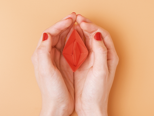 Mãos femininas com unhas pintadas de vermelho seguram uma representação estilizada de uma vulva vermelha em fundo laranja claro.